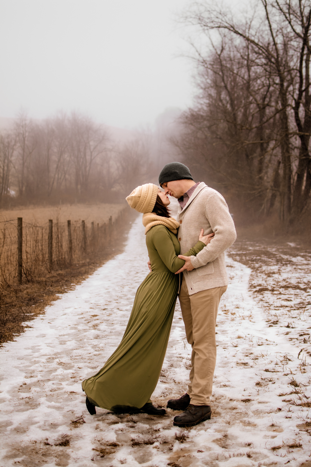 Winter adventure session attire in the Hocking Hills. A couple coordinates in warm earth tones and rich textures, featuring a woman in a sage green dress and cream scarf, and a man in a cream fisherman cardigan, on a snowy, foggy trail.