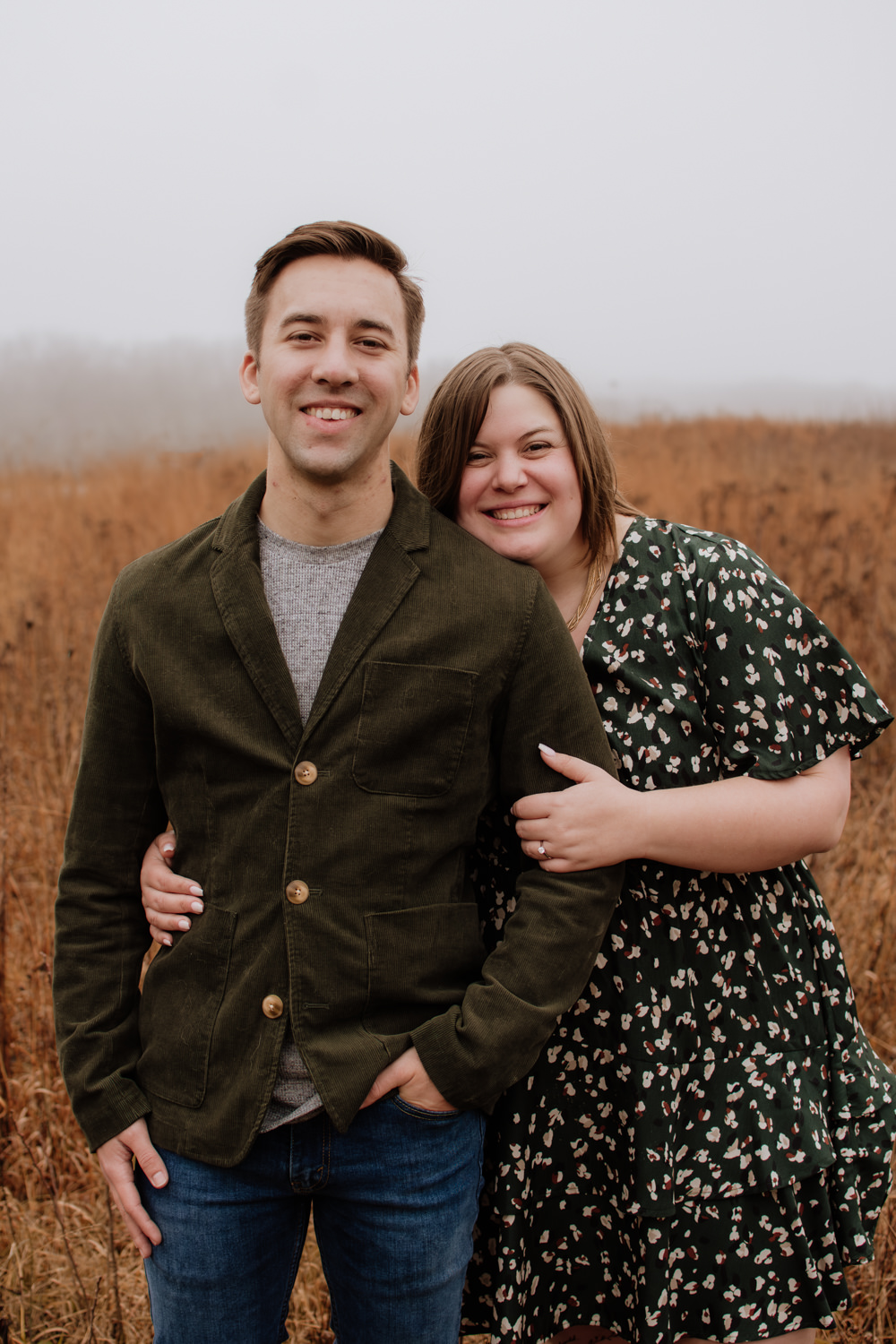 Couple wardrobe inspiration for a Hocking Hills trail session. The man wears a textured deep green corduroy blazer and denim, while the woman wears a flowing green floral dress, demonstrating how to mix rich textures and movement against a natural landscape.