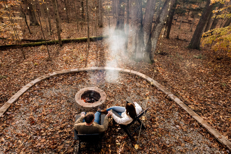 High-angle shot of the couple sitting together outside The Pine cabin, with the brilliant orange and yellow November sunset filtering through the tree canopy behind them. The fire in the pit smokes in the evening light.