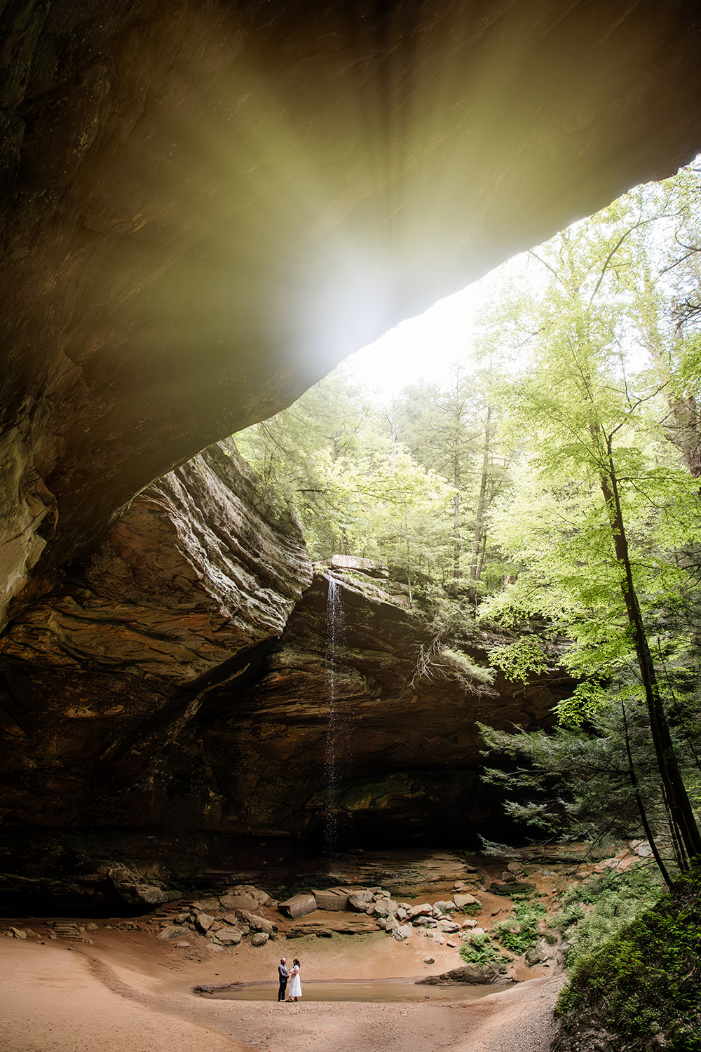 A couple stands face-to-face inside the vast sandstone recess of Ash Cave in Hocking Hills, exchanging private wedding vows. The towering cave walls frame their intimate ceremony, highlighting the quiet connection between them in the massive natural cathedral.
