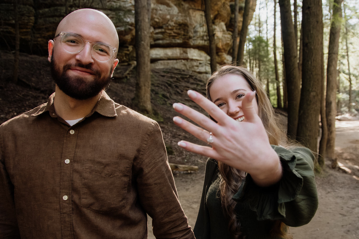 A close-up of an engaged couple in a wooded setting, with the woman’s hand held toward the camera to showcase a sparkling engagement ring. The couple is softly blurred in the background, focusing attention on the ring and the texture of the forest environment.