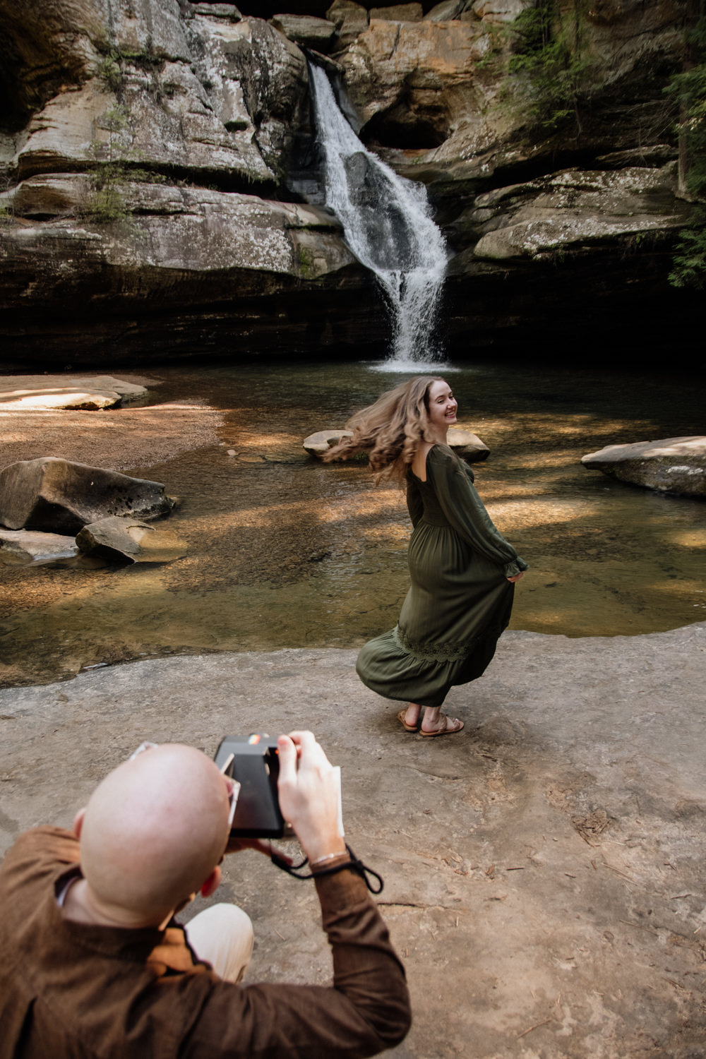 A couple stands before the rushing white water of Cedar Falls; the woman twirls in a long, mossy green dress that catches the light, while the man leans in to capture her movement with a vintage Polaroid camera.