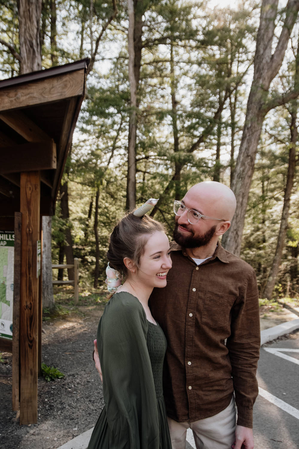 An engaged couple arrives at the Cedar Falls trailhead in the Hocking Hills; the woman wears a light-colored heatless curling rod wrapped over the top of her head, resembling a unicorn horn, as they prepare for their spring session.