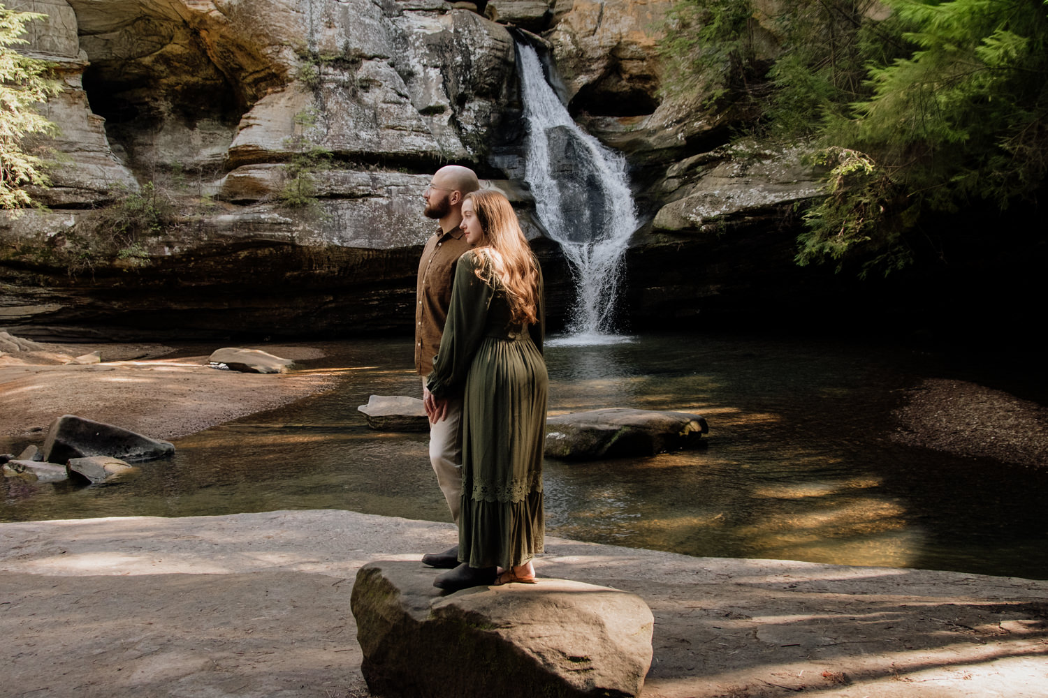 An engaged couple stands poised against a towering, weathered rock formation; the woman rests her head gently on the man’s shoulder with a serene expression, their steady and graceful pose resembling a classical stone sculpture amidst the rugged textures of the gorge.