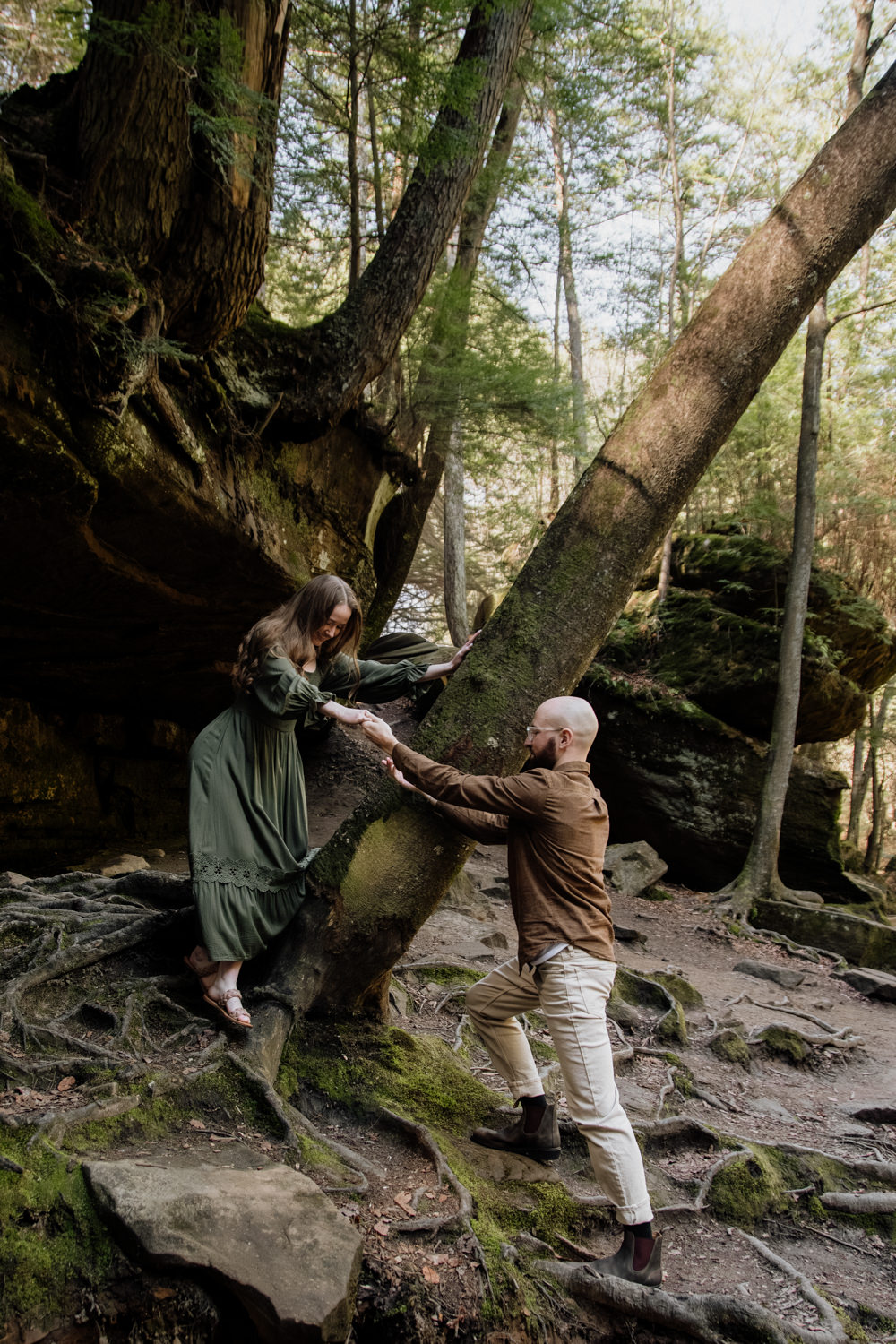 A man reaches out his hand to support a woman as she carefully steps down from a moss-covered rock and tree trunk, capturing a supportive and gentle moment in a lush forest of Hocking Hills.