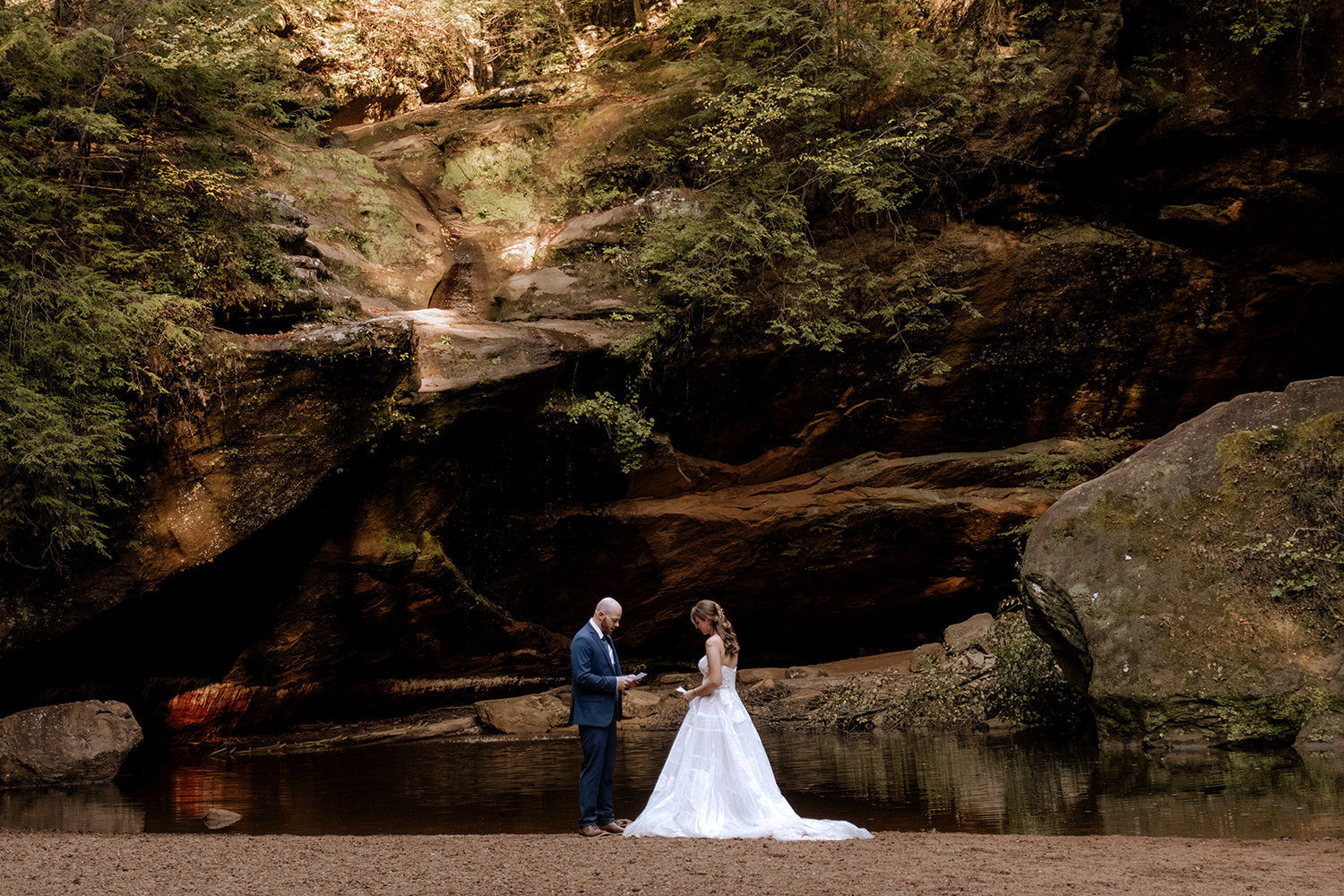 An intimate moment of a couple reading elopement vows at Lower Falls in Hocking Hills, Ohio. The scene is surrounded by fall foliage and the natural stone textures of the gorge.