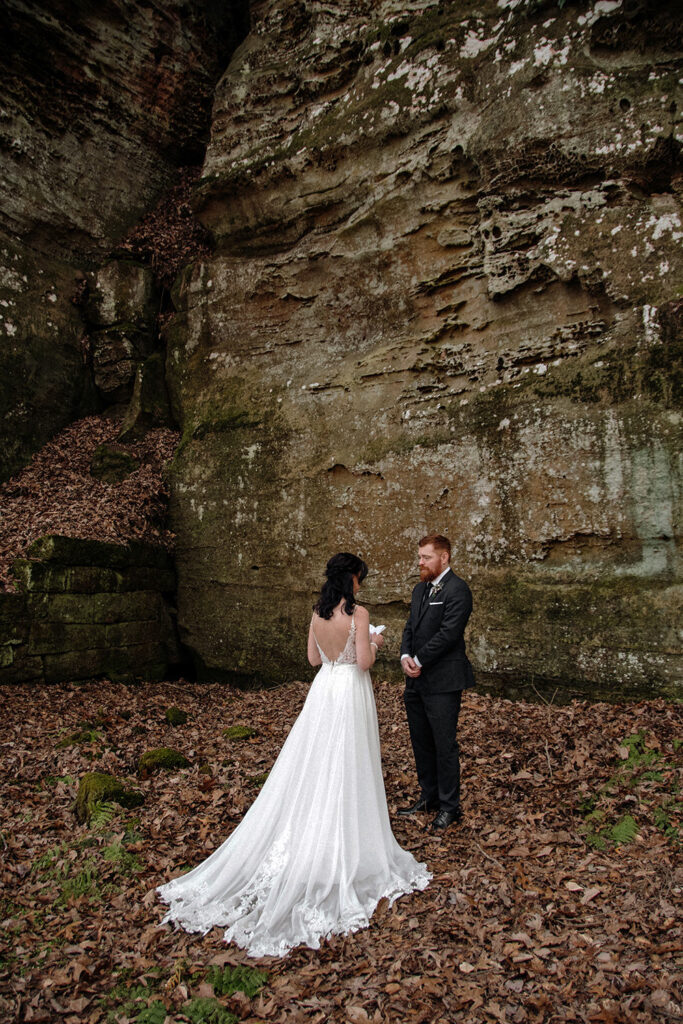 An intimate elopement scene of a couple reading vows to one another while standing at the base of a massive sandstone cliff. The secluded location emphasizes the privacy and intentionality of their ceremony.