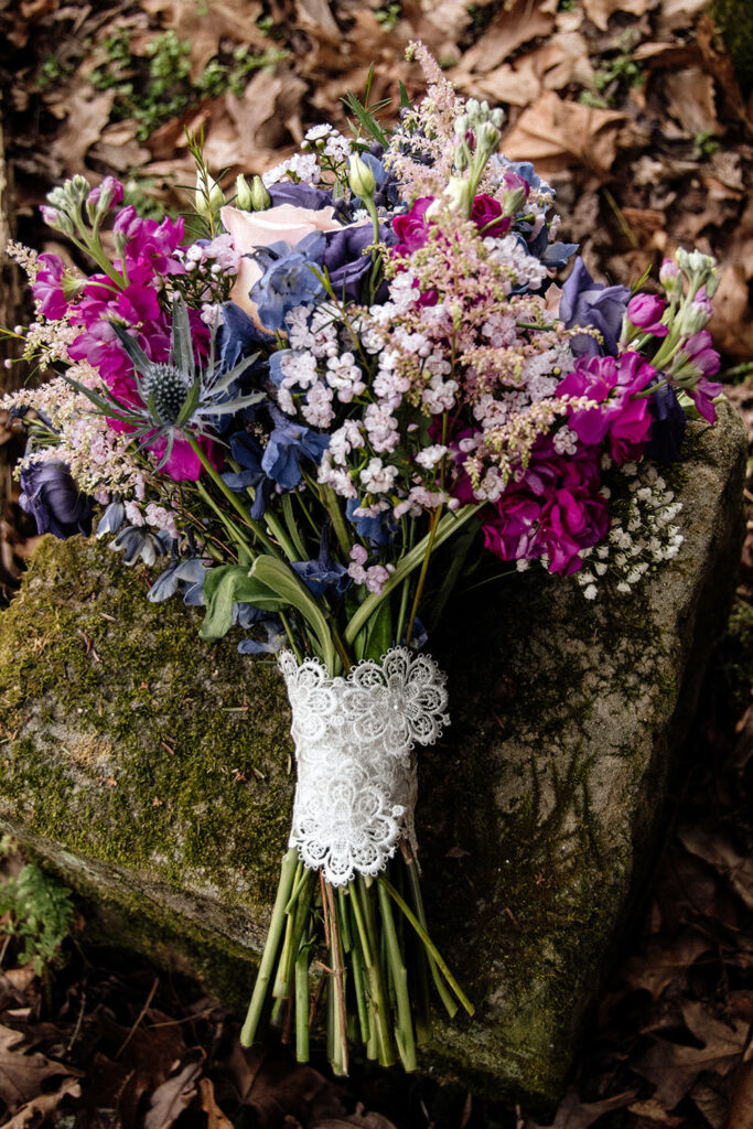 A bridal wedding bouquet featuring a mix of pink, purple, and white flowers resting on a textured green rock. The stems are elegantly bound with delicate white lace.