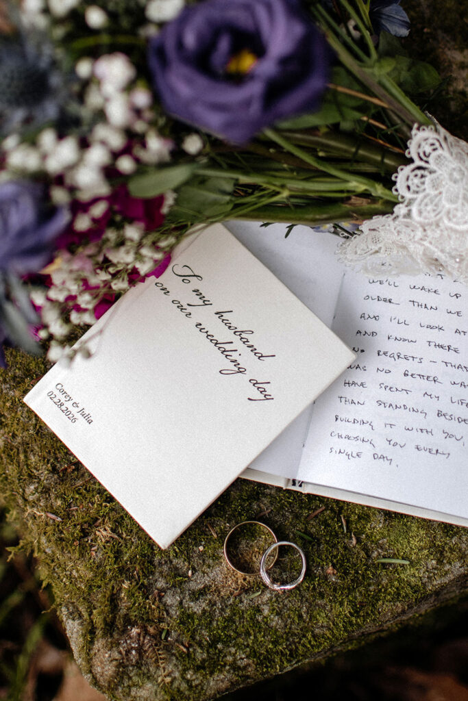 Two elopement vow books rest on a lush, mossy rock in a forest setting. One book is open, revealing handwritten notes alongside two wedding rings and a bridal bouquet. The scene captures the intimate, intentional details of a private ceremony.