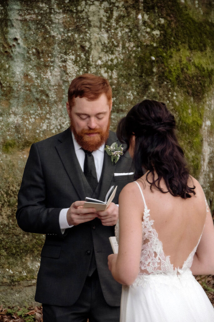 A red-haired groom stands in a secluded, rocky alcove in Hocking Hills, reading from a vow book to his bride. The intimate elopement scene is tucked away in a hidden forest location, captured with a focus on the couple's quiet connection.