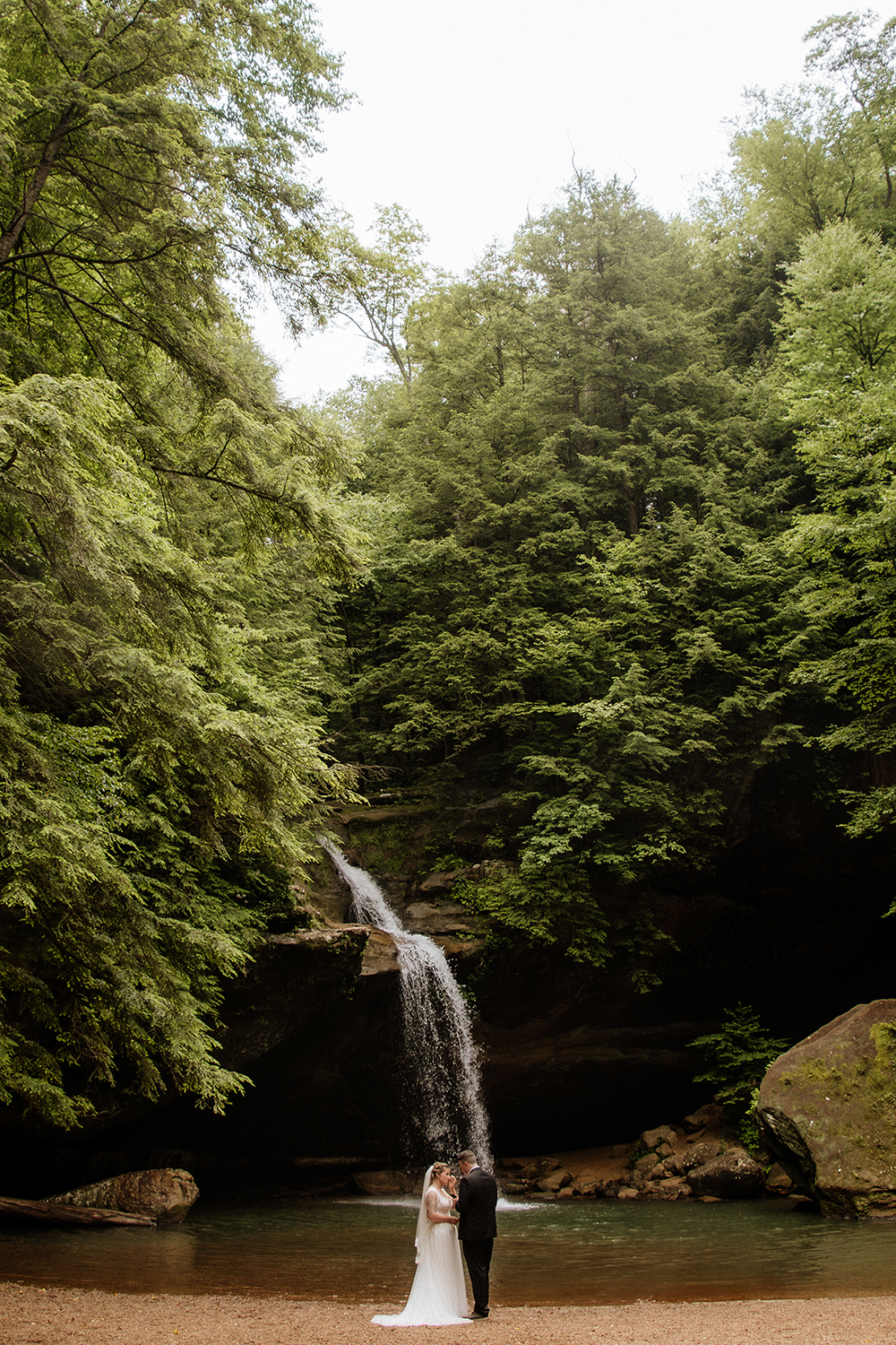 A wide shot of a couple exchanging private wedding vows at the base of Lower Falls in Old Man’s Cave, surrounded by the iconic rock formations and forest of Hocking Hills.