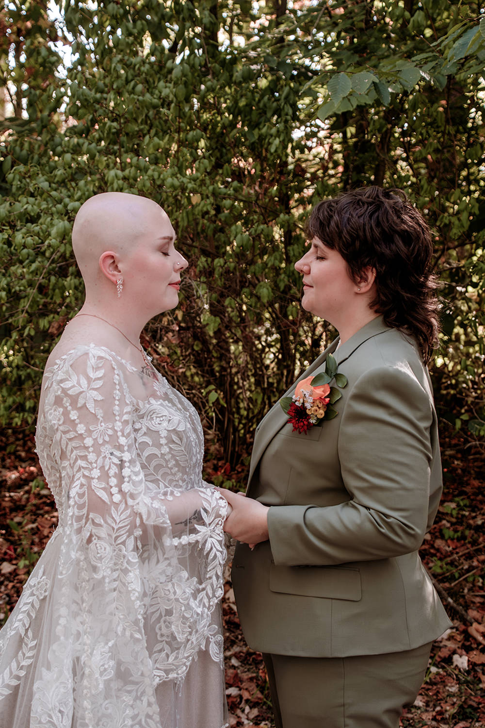 Option 1 (Descriptive & Detailed):
A queer couple stands close together with their eyes closed, holding hands during their private wedding vows. One woman, with a shaved head, wears a white boho-style wedding dress, while the other woman wears a tailored green suit.