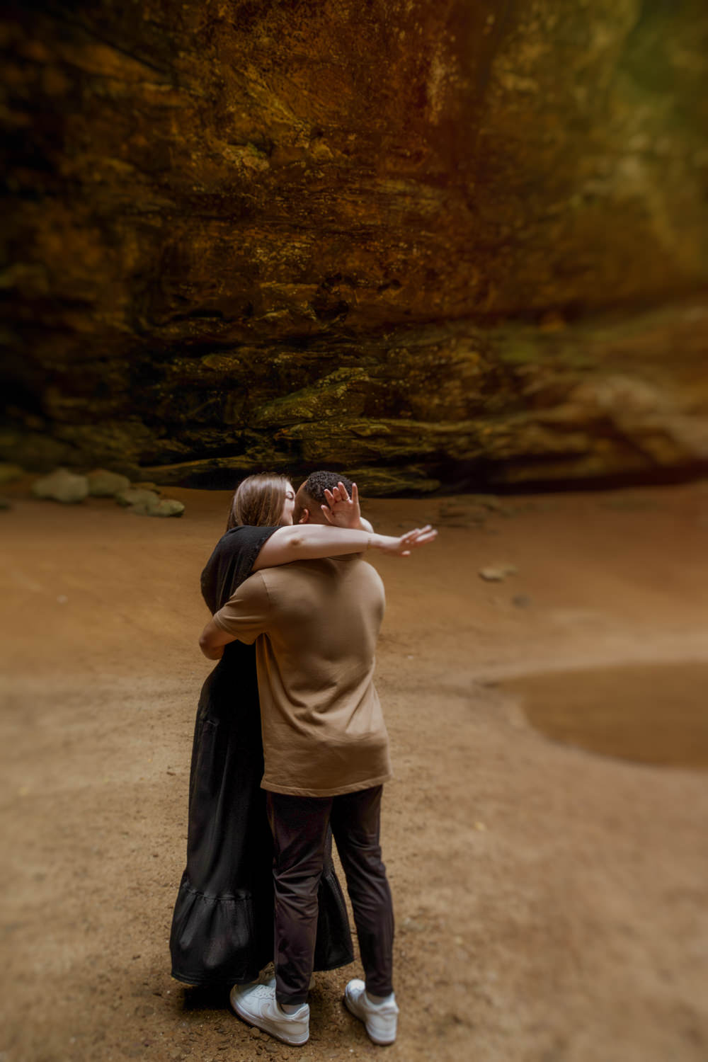 A man and woman embrace by the waterfall pool at Ash Cave. The epic wall of the tall cave is behind them.