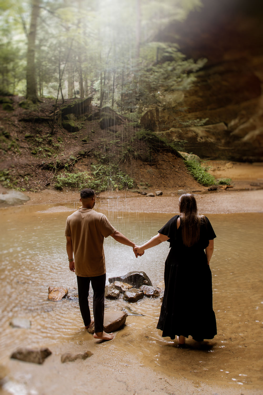 A man and woman hold hands and stand beneath a waterfall in the Hocking Hills.
