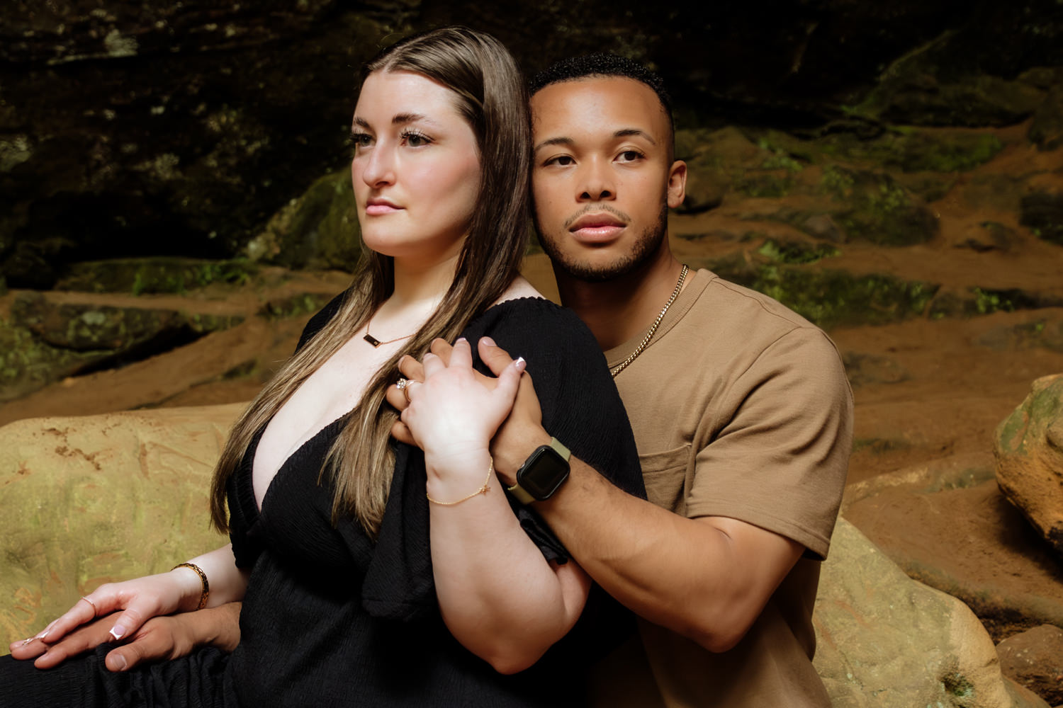 A black man holds a white woman's hand and they look in opposite directions. The woman is wearing a unique engagement ring on her finger.