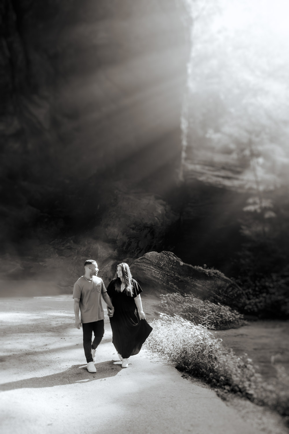 A man and woman hold hands and walk away from Ash Cave as rays of light form behind them.