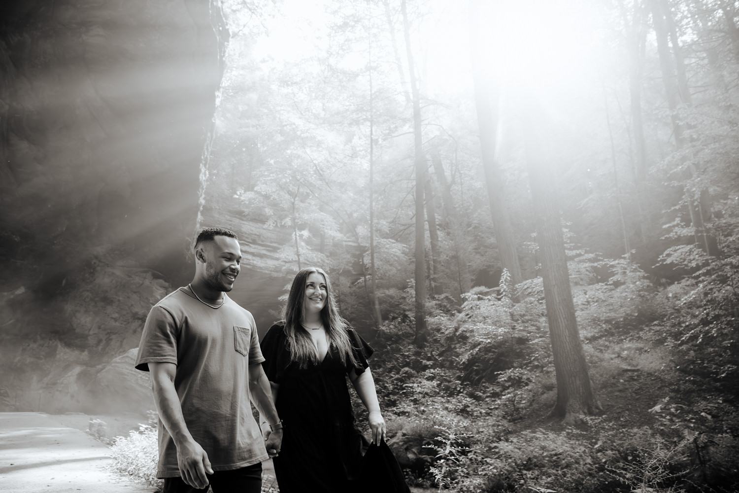 Black and white photo of a couple holding hands and walking out of Ash Cave with sun rays behind them.