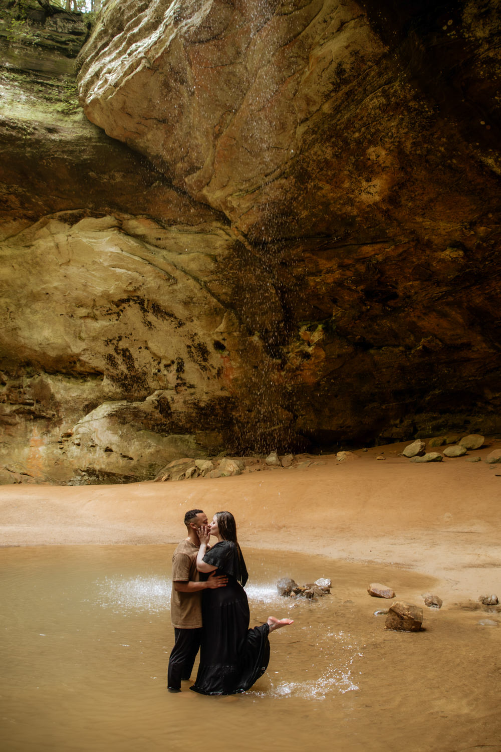 A man and woman kiss beneath a waterfall in Hocking Hills, Ohio. The woman lifts her bare foot and points it up.