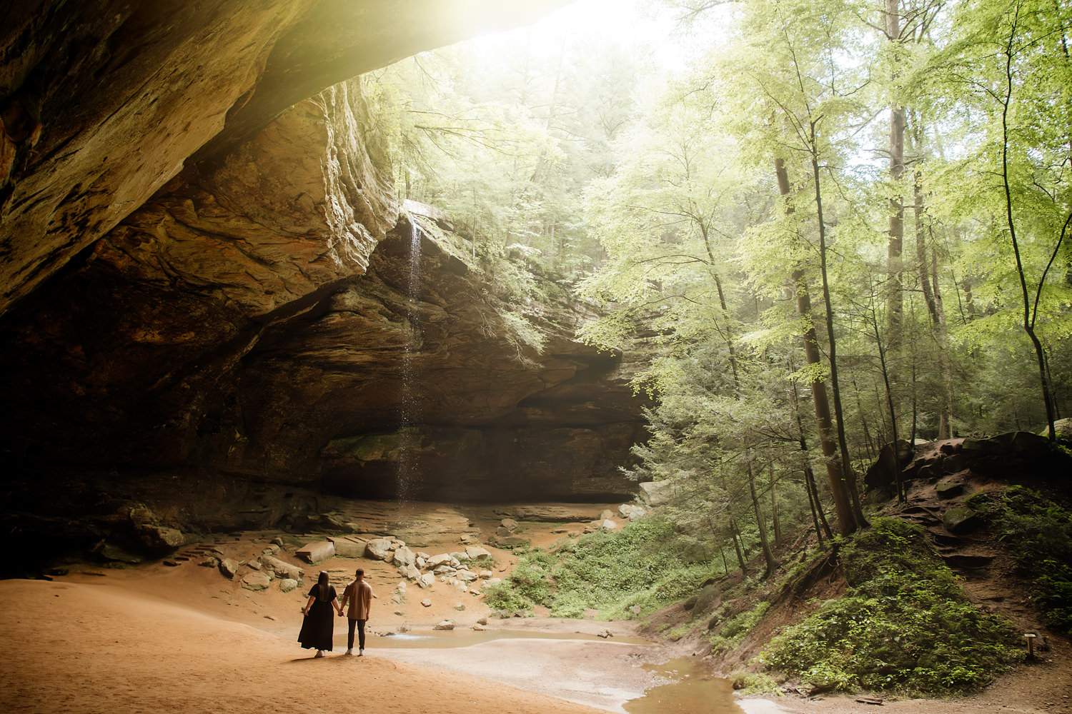 A wide view of Ash Cave with a light flare at the top of the cave rim and a tiny couple at the bottom holding hands.