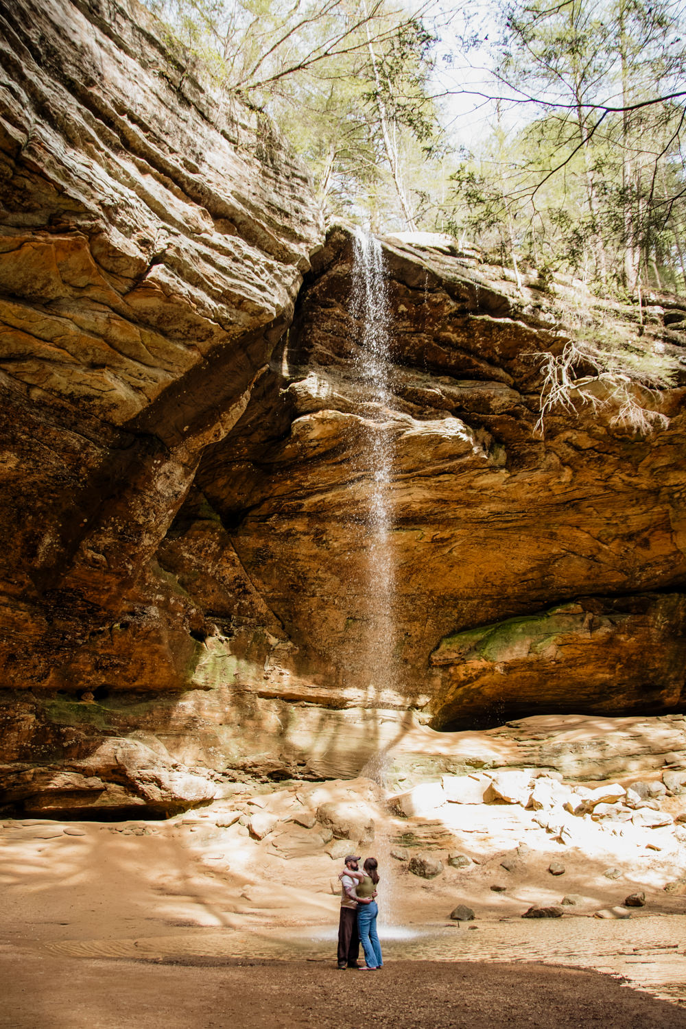 A couples stands under the waterfall at Ash Cave. Golden light surrounds them.