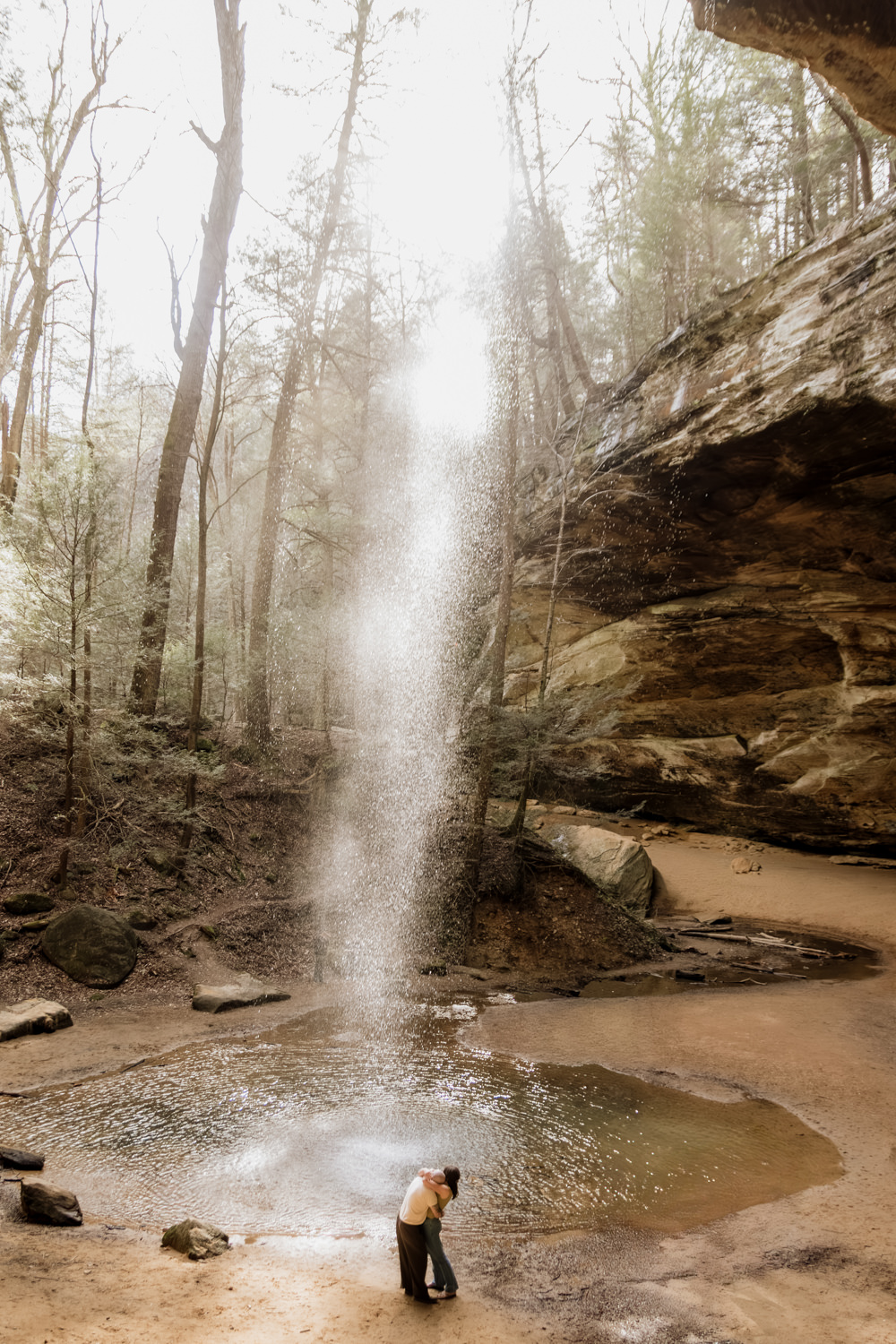 A wide vertical shot of the waterfall at Ash Cave with a couple standing beneath it.