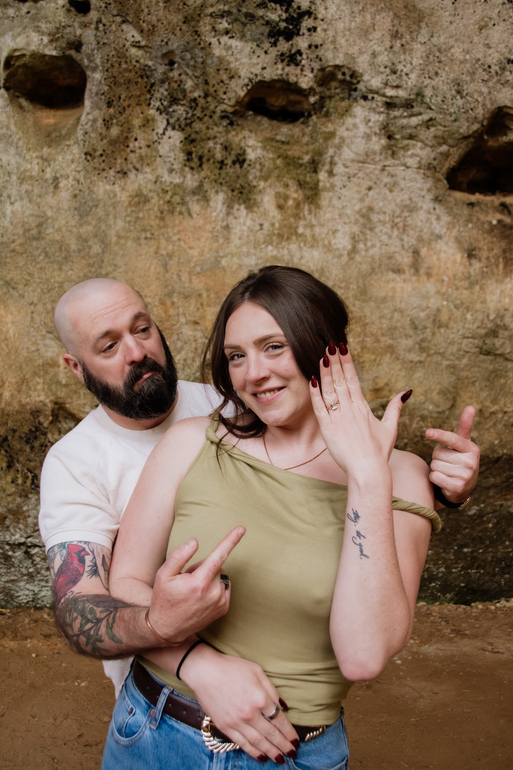A silly candid couple photo taken after a surprise proposal. The man points to the engagement ring and the woman holds her ring hand towards the camera.