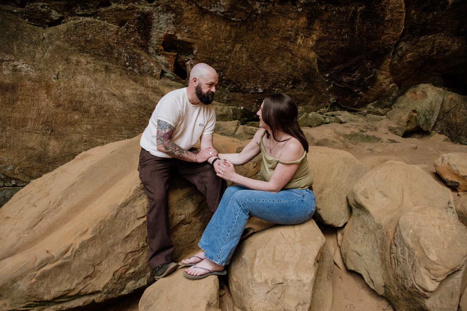 A man and woman hold hands and lean towards each other while sitting on rocks at Ash Cave.