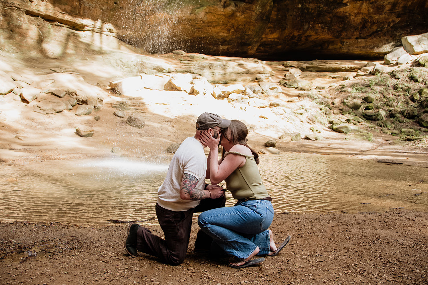A man and woman crouch on their knees after he proposed to her by the waterfall at Ash Cave.