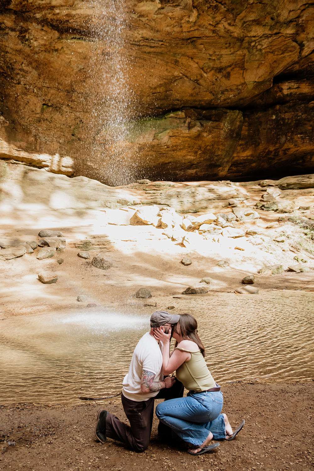 A man and woman crouch on their knees after he proposed to her by the waterfall at Ash Cave.
