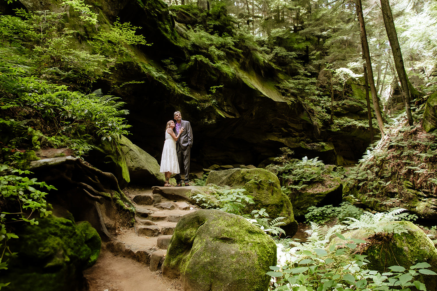 A couple celebrates their 25th wedding anniversary by getting dressed up and hiking to a special spot in Conkle's Hollow Gorge for a photo session. The couple appears tiny and dwarfed by the green ferns and moss of the gorge.