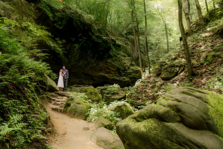A couple stands in Conkles Hollow gorge for their 25th anniversary portraits. They are dwarfed by rock walls and vibrant green.
