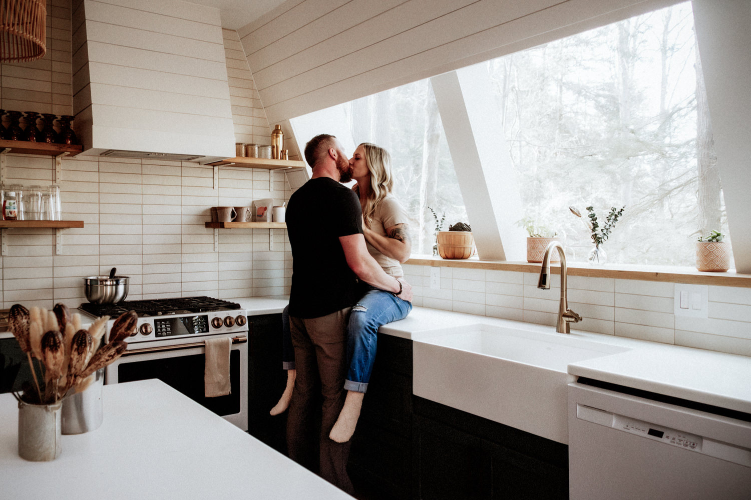 A woman sits on a counter in the modern open plan kitchen of Dunlap Hollow A-frame. The man leans in and kisses her on the lips.