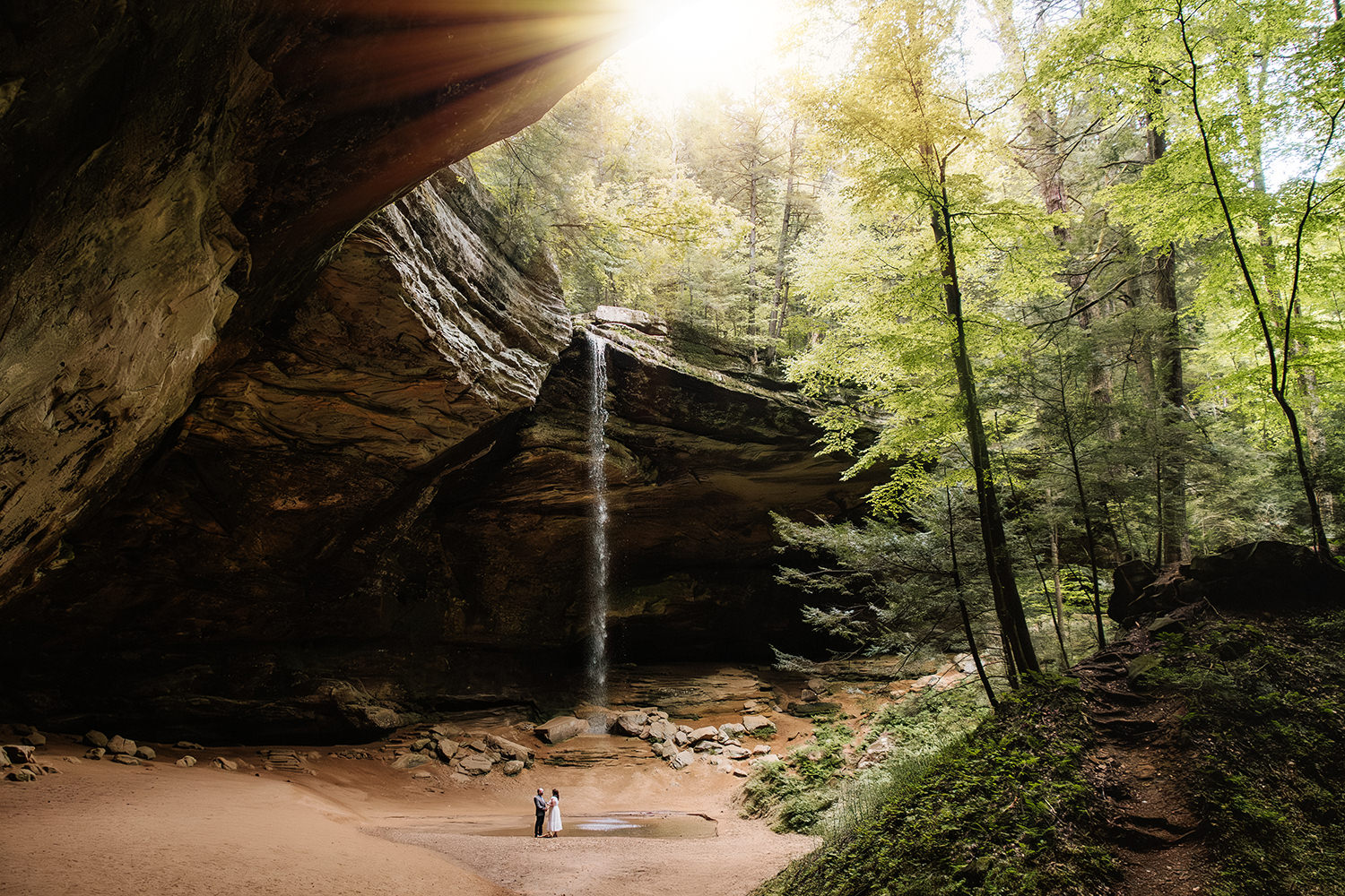 An elopement couple is dwarfed by the horseshoe-shaped overhang of Ash Cave in the Hocking Hills, Ohio.