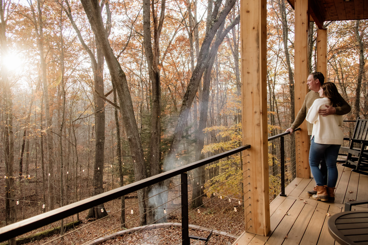 Wide shot of Taylor and Jeff standing on the spacious wooden deck of The Pine at Dunlap Ridge cabin, surrounded by towering Hocking Hills pine trees glowing in the November golden hour light.