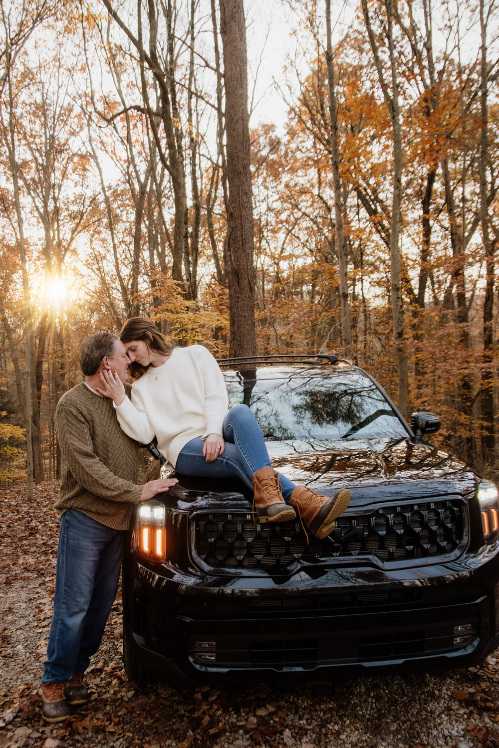 A sweet candid photo of Jeff kissing Taylor’s forehead while they stand and sit on a black SUV near The Pine cabin, surrounded by autumn colors.