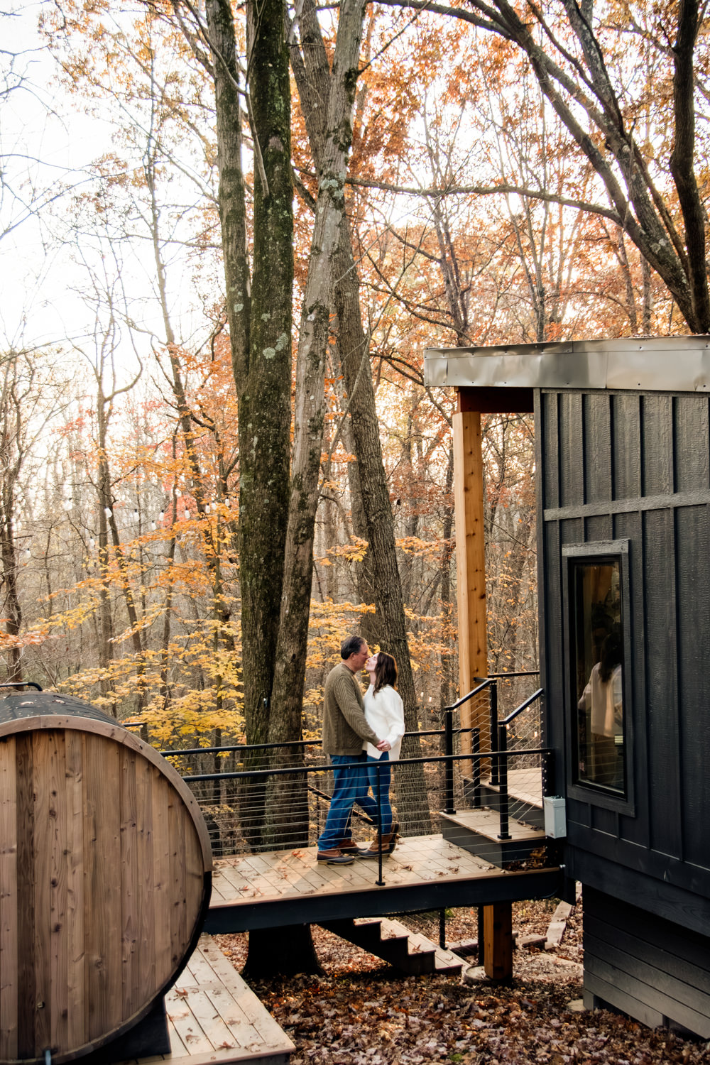 An exterior view of The Pine, a modern black A-frame cabin, with Taylor and Jeff holding hands on the front porch during a warm, hazy autumn sunset.