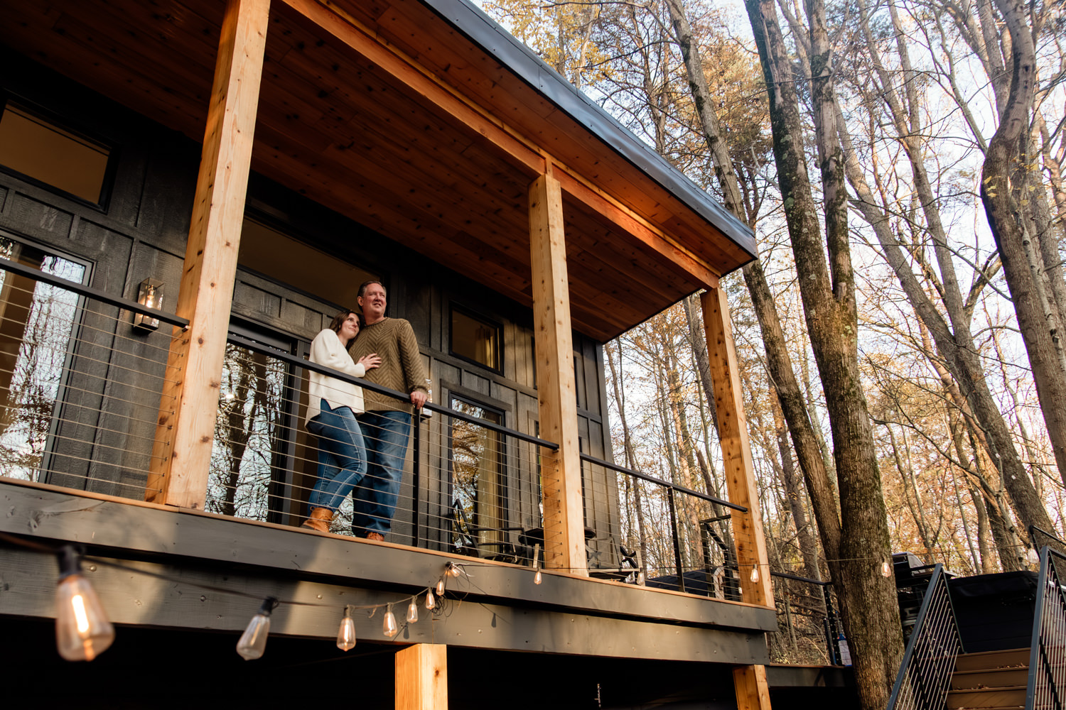 Wide shot of Taylor and Jeff standing on the spacious wooden deck of The Pine at Dunlap Ridge cabin, surrounded by towering Hocking Hills pine trees glowing in the November golden hour light.