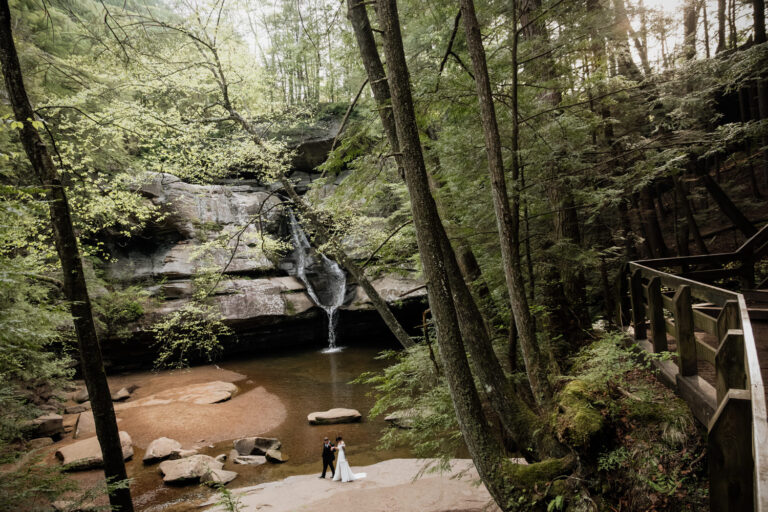 A wide shot of Cedar Falls at Hocking Hills State Park. A tiny elopement couple walks by in the mid distance.