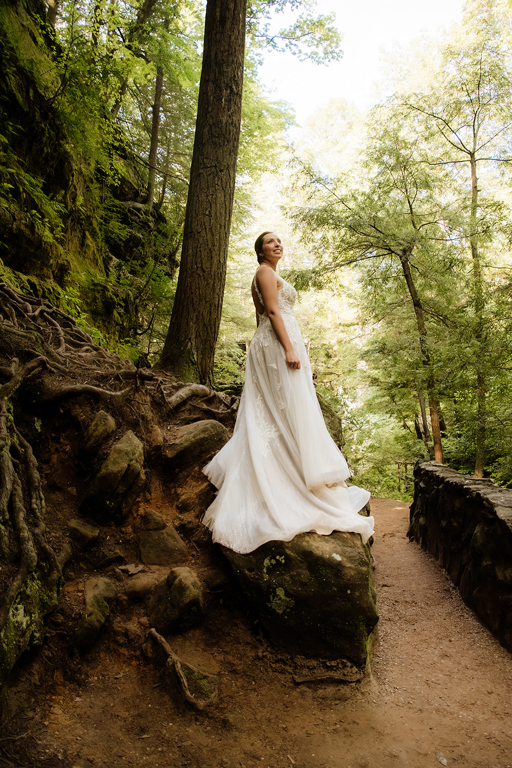 A bride stands on a mossy rock with tree roots. She twists toward the camera and the forest light is magical behind her.