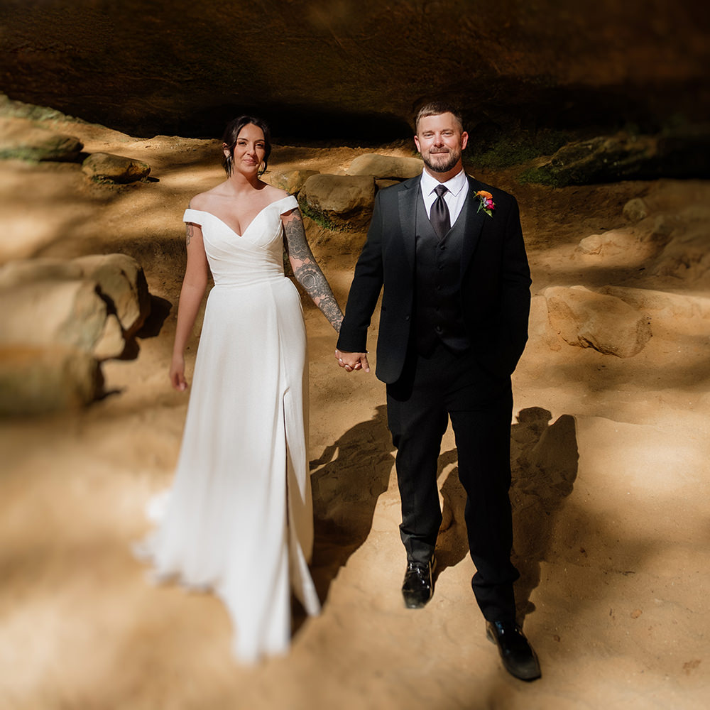 A bride in off the shoulder modern dress with vintage styling walks and holds hands with a groom in a classic black suit.  The couple is photographed inside the rocky and sandy area of Ash Cave in in the Hocking Hills, Ohio.