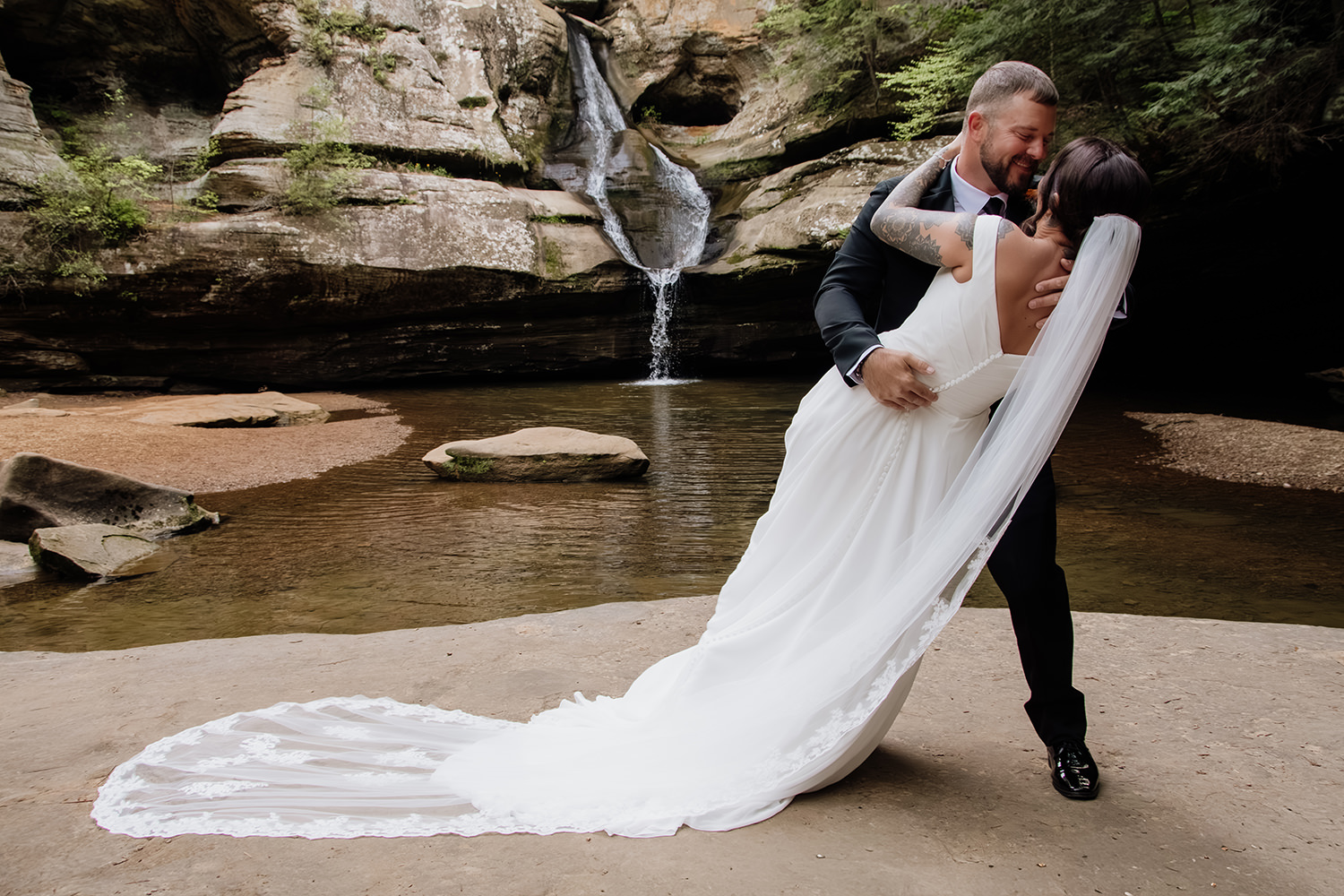 A man dips a woman into a dramatic kiss at their Cedar Falls elopement.  A waterfall flows in the background of this Hocking Hills, Ohio location.