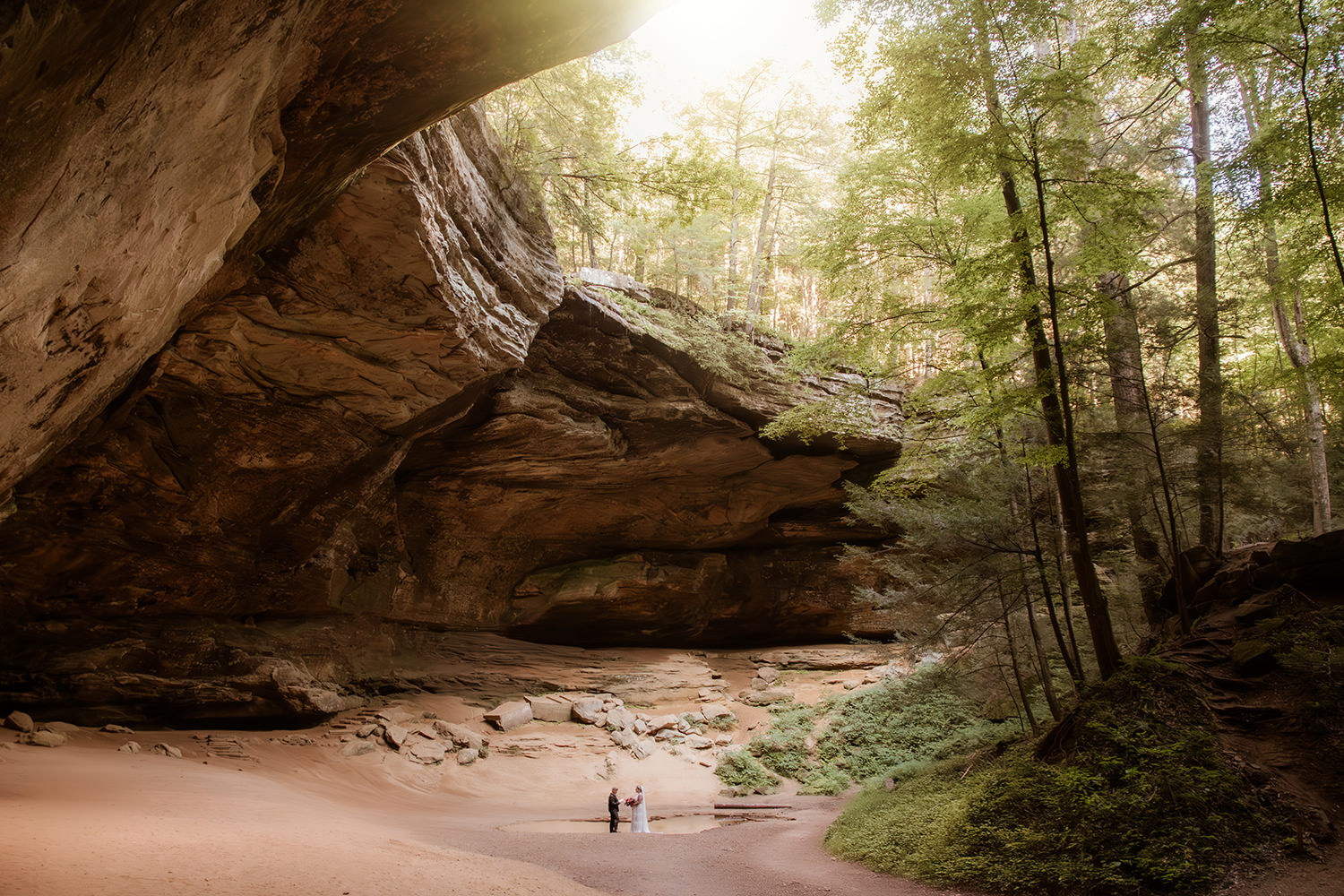 The epic rim of Ash Cave hangs in the light as a lesbian couple says private vows to each other at the base.  The couple looks tiny next to the towering scale of the cave.