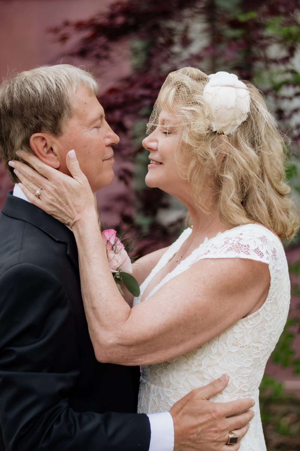 An older couple kisses in front of a pink wall with bright green foliage at Ravenwood Castle in the Hocking Hills.