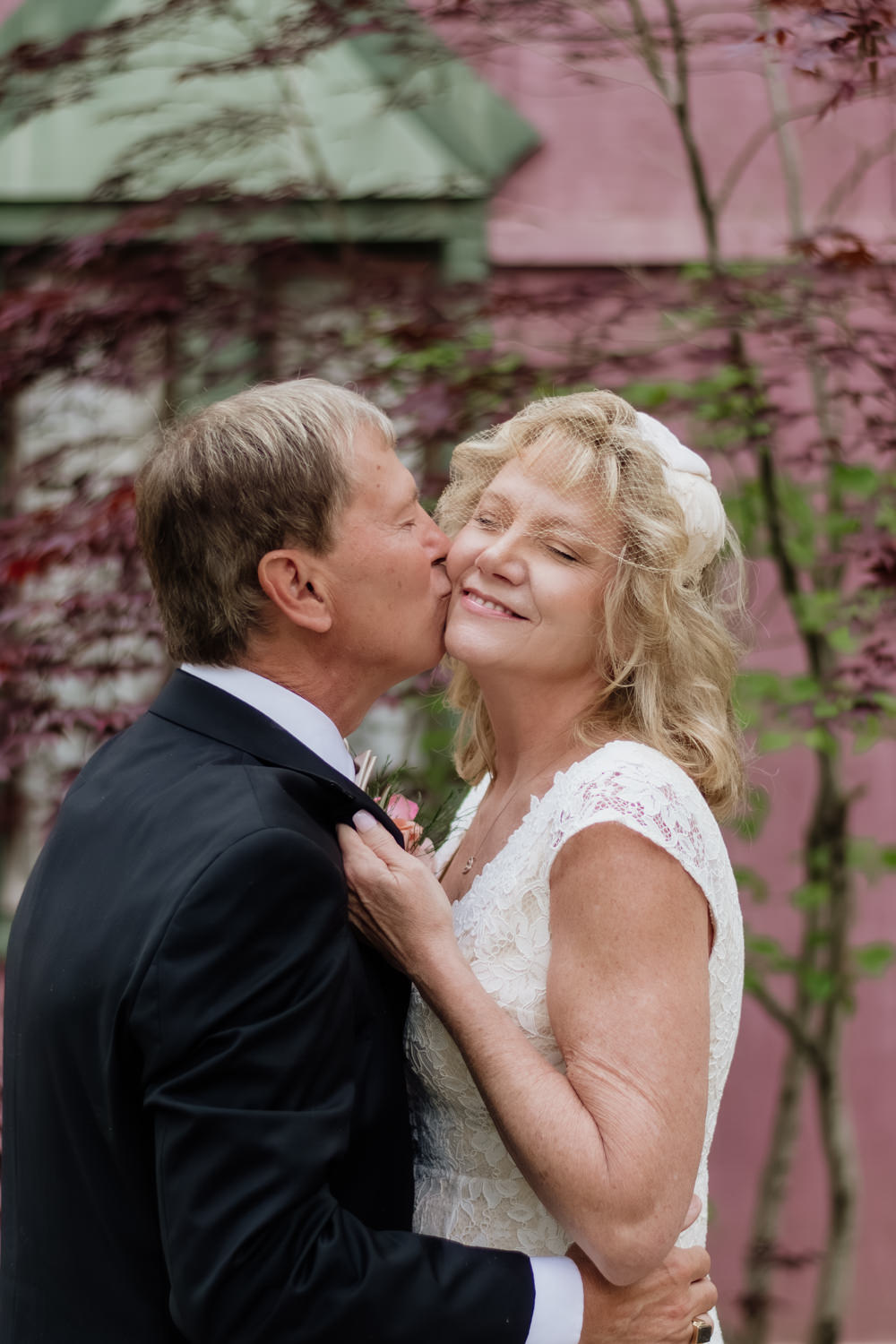 An older couple kisses in front of a pink wall with bright green foliage at Ravenwood Castle in the Hocking Hills.