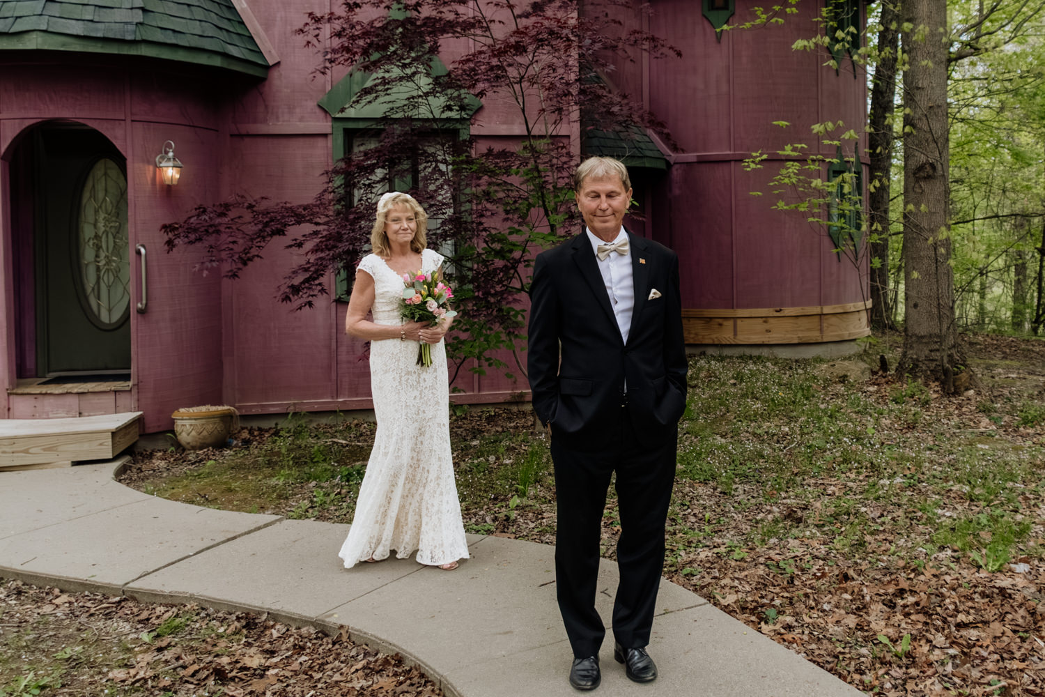 A woman approaches a man from behind. The man looks at the camera. In the background is the pink Silversmith's Cottage at Ravenwood Castle.