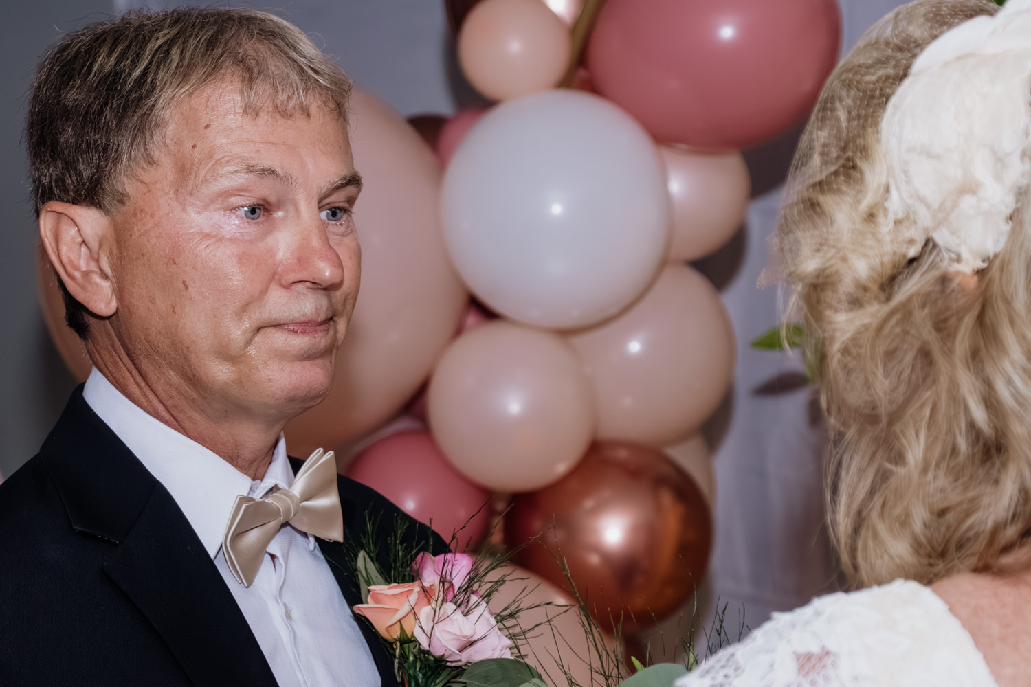 A teary eyed man recites his vows during his 35th anniversary renewal. In the background is a string of rose colored balloons.