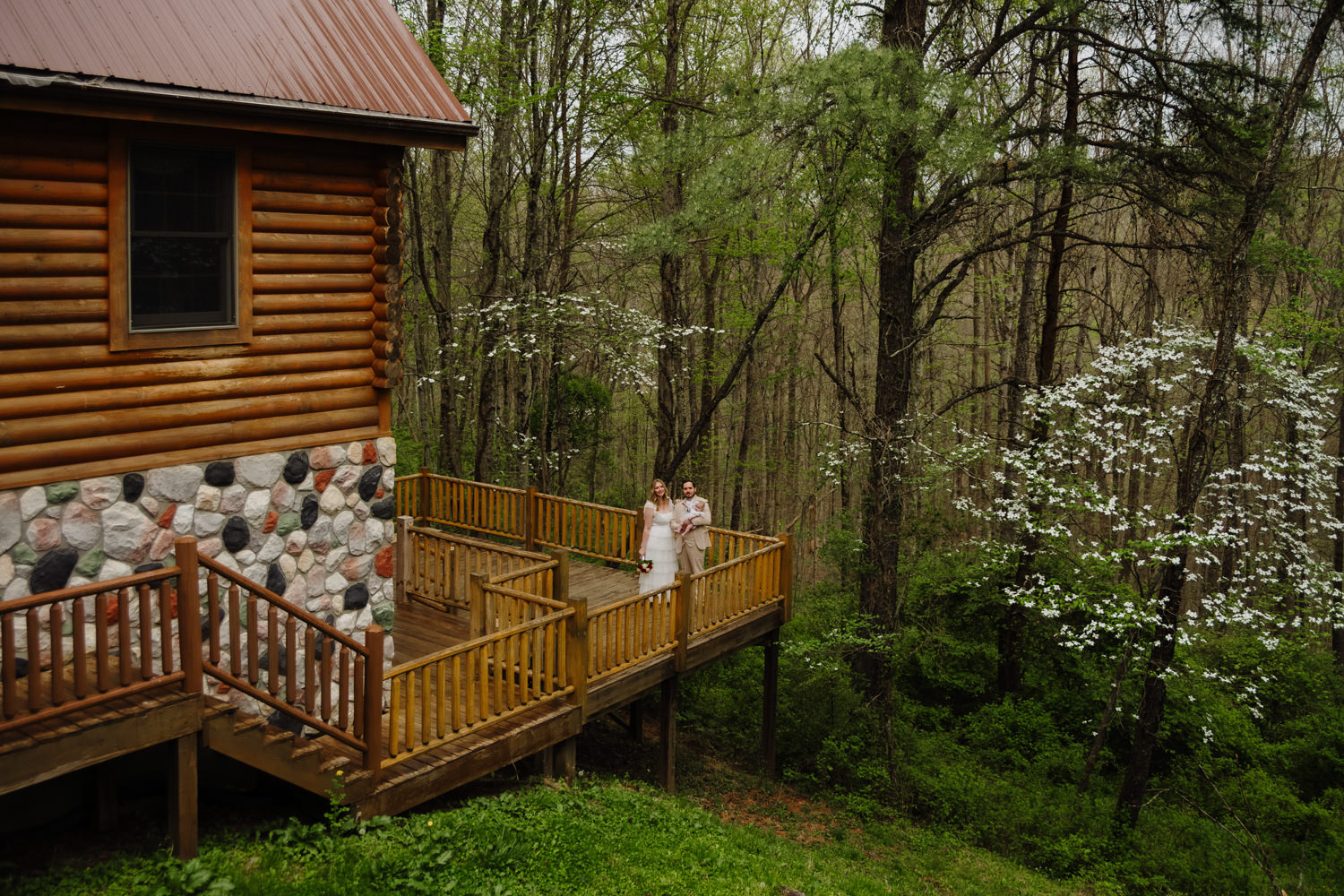 A couple holds a baby and stands next to a stone fireplace and a wooden pergola at Hocking Hills Wedding Chapel in spring.  The couple renews their vows on their 5th wedding anniversary.