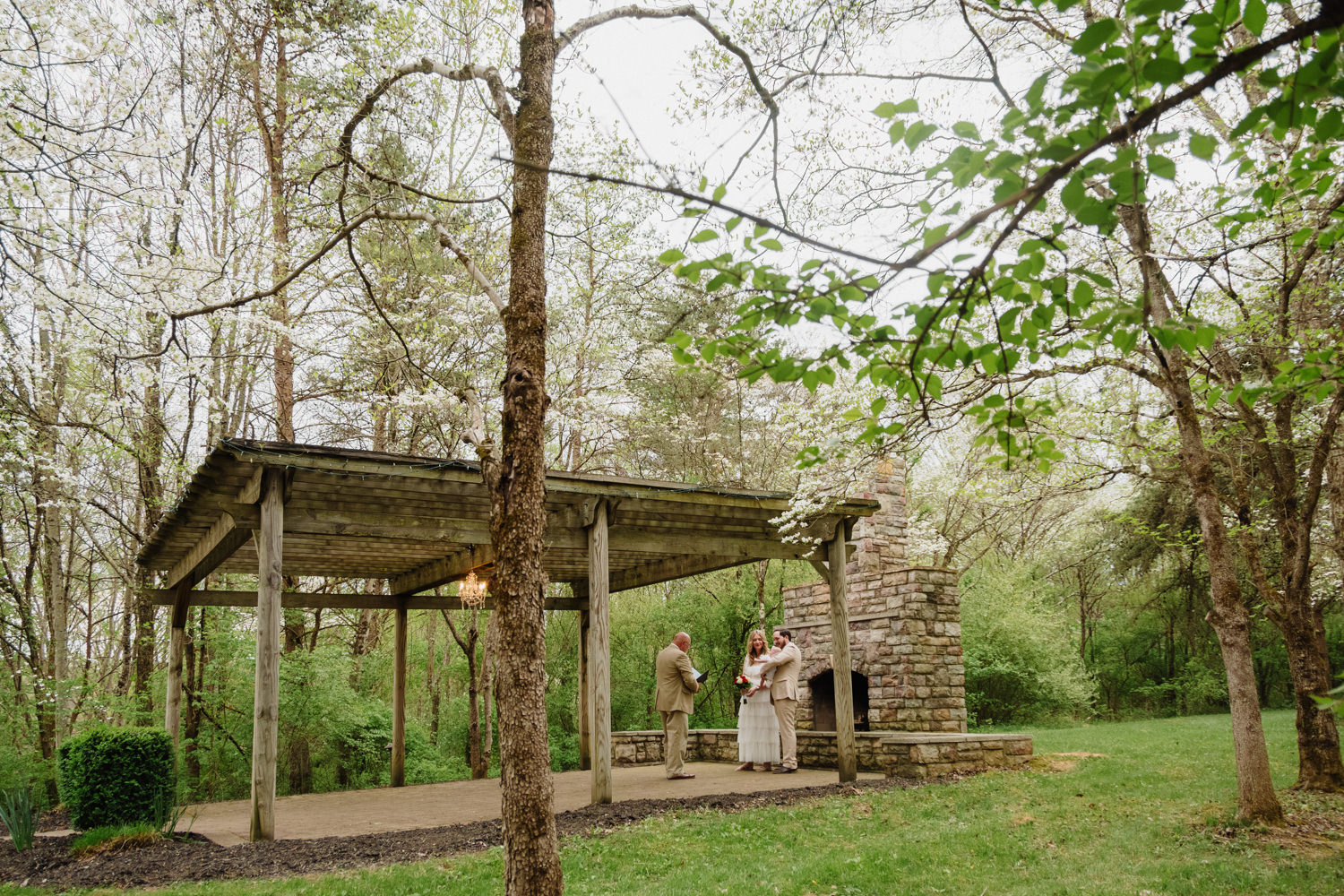 A couple holds a baby and stands next to a stone fireplace and a wooden pergola at Hocking Hills Wedding Chapel in spring. The couple renews their vows on their 5th wedding anniversary.