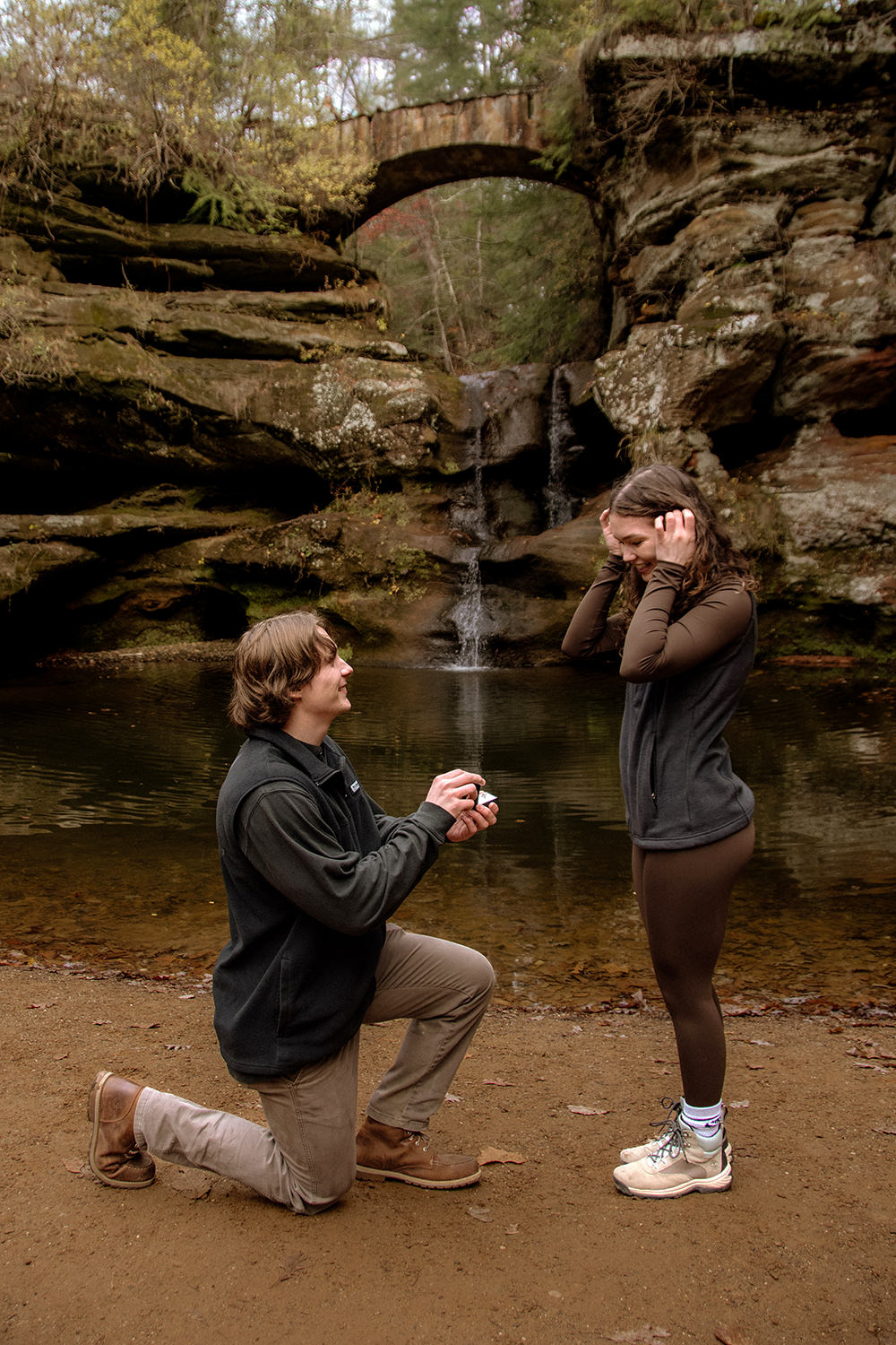 A man on one knee proposes to a woman in front of Upper Falls at Old Man's Cave during November. There are surrounded by brown fall tones.