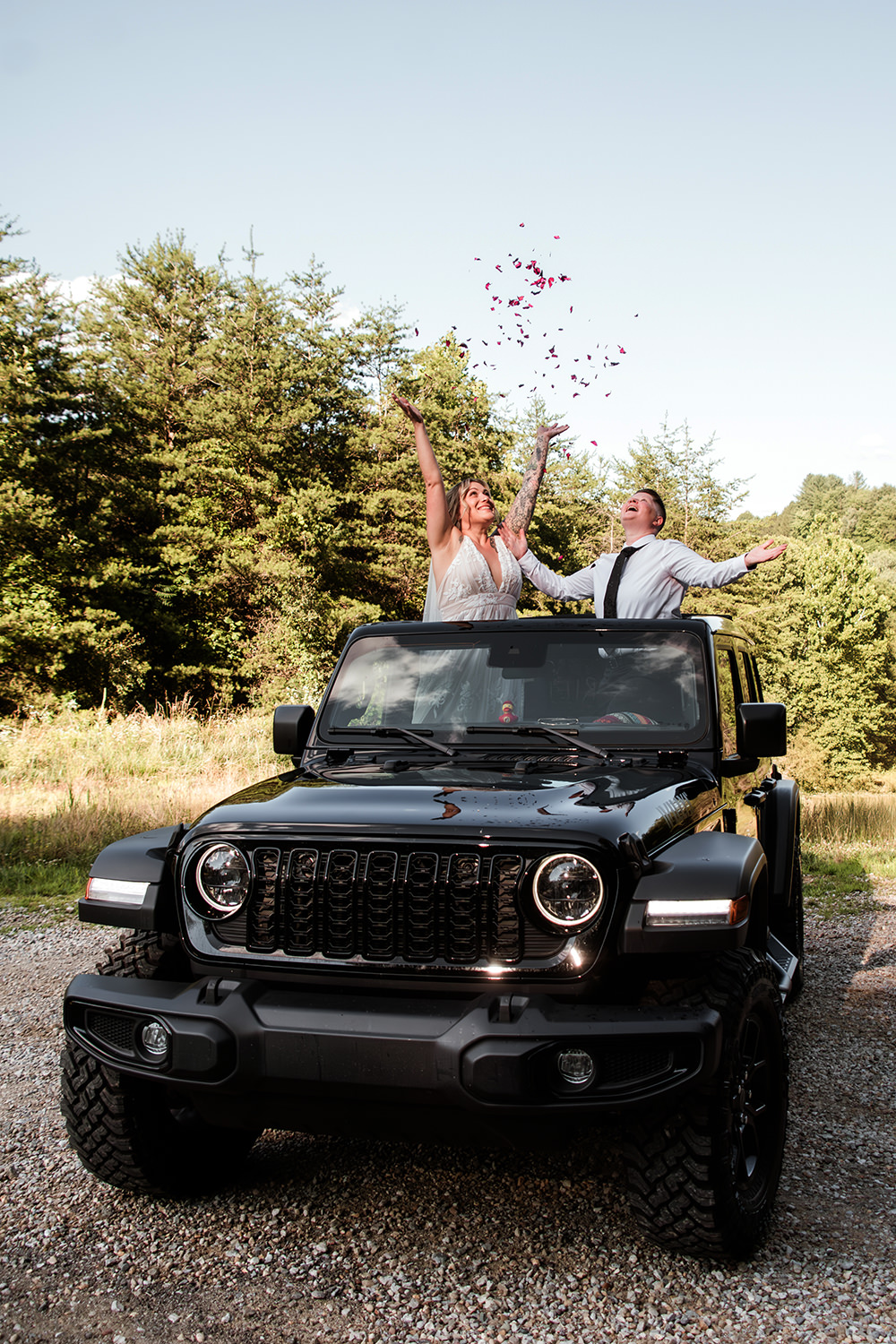 A queer couple stands through the sun roof of a jeep and they throw confetti up in the air.